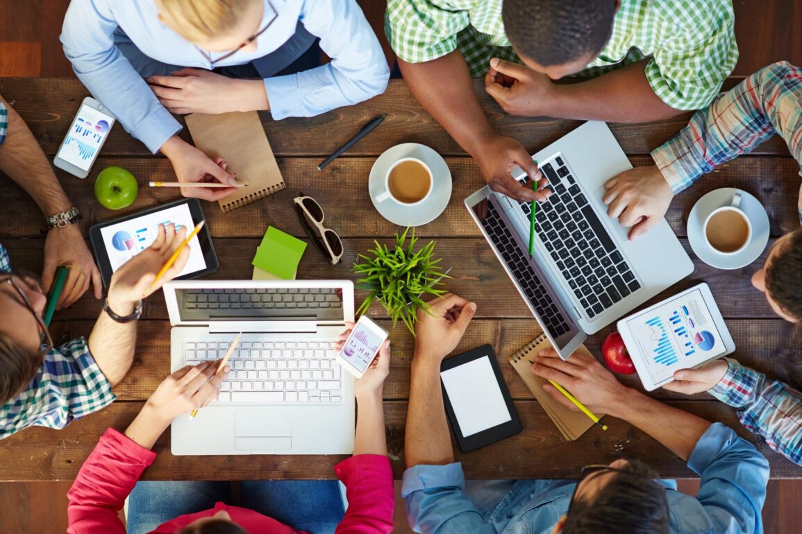 Top-down view of people communicating at table with their devices and notepads.